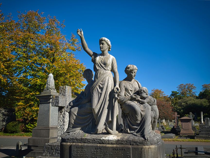 A paved pathway in Brompton Cemetery lined with lush green trees on both sides leads towards a historic domed mausoleum structure in the distance. The surface of the pathway appears clean and well-mai