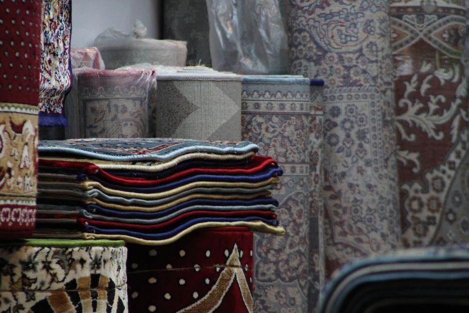 A neatly stacked arrangement of various folded carpets and rugs with intricate patterns and vibrant colors, including reds, blues, and creams, displayed on a surface in a well-lit room. The background