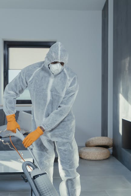 A professional cleaner dressed in white protective disposable coveralls, orange gloves, and a face mask is operating a vacuum cleaner on a clean, modern residential floor. The room features large wind